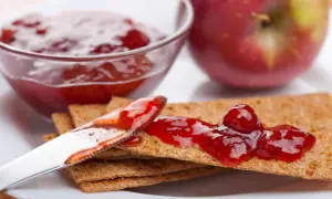 Un cucchiaio spalma la marmellata di mele su cracker croccanti, con una ciotola piena e una mela rossa sullo sfondo.
