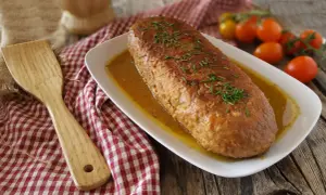 Polpettone in padella dorato e succulento, guarnito con erba cipollina, su un piatto bianco con salsa. Accanto, una spatola di legno e pomodorini.