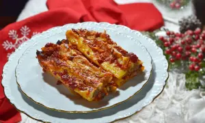 Due cannelloni di carne fumanti, ricoperti di sugo rosso e formaggio fuso, serviti su un piatto bianco con decorazioni natalizie sullo sfondo.