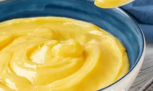 Una ciotola blu piena di vellutata crema pasticcera senza uova, con un cucchiaio che ne solleva una porzione.