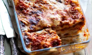 Lasagne alla bolognese appena sfornate, dorate e filanti, in una teglia di vetro. Accanto, due coltelli d'argento.
