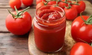 Un vasetto di ketchup fatto in casa, denso e rosso vivo, circondato da pomodori freschi su un tagliere di legno.