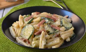 Piatto di pasta cremosa con zucchine a rondelle e cubetti di speck croccante, guarnito con pepe nero.