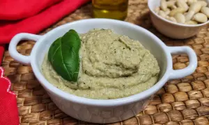 Pesto di melanzane cremoso in una ciotola bianca, guarnito con una foglia di basilico. Sullo sfondo, mandorle e olio d'oliva.
