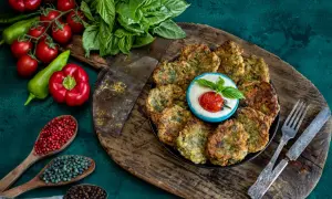 Piatto di polpette di zucchine dorate, servite con una ciotolina di salsa bianca, pomodorino e basilico. Ingredienti freschi e spezie completano la scena.