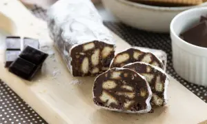 Un delizioso salame al cioccolato, cosparso di zucchero a velo, con biscotti e cioccolato fondente a lato.