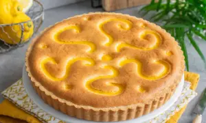 Torta con crema pasticcera, decorata con un motivo a spirale giallo brillante. Sullo sfondo, limoni freschi e foglie verdi.