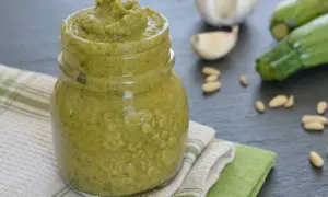 Un vasetto pieno di cremoso pesto di zucchine, con ingredienti freschi come zucchine, aglio e pinoli sullo sfondo.