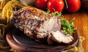 Arista di maiale succulenta e dorata, affettata su un tagliere di legno, con rosmarino e pomodori sullo sfondo.