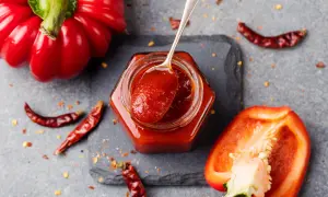 Un vasetto di marmellata di peperoni, con un cucchiaino che ne raccoglie un po'. Peperoni rossi interi e tagliati, peperoncini secchi e fiocchi di peperoncino completano la scena.