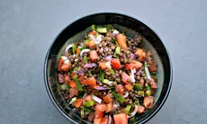 Una fresca insalata di lenticchie estiva, con pomodori, cipolla rossa e erbe aromatiche, servita in una ciotola scura.