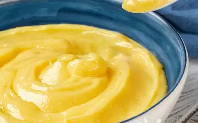 Una ciotola blu piena di vellutata crema pasticcera senza uova, con un cucchiaio che ne solleva una porzione.