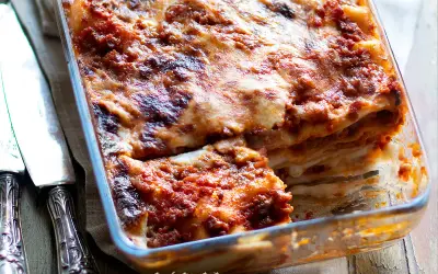 Lasagne alla bolognese appena sfornate, dorate e filanti, in una teglia di vetro. Accanto, due coltelli d'argento.