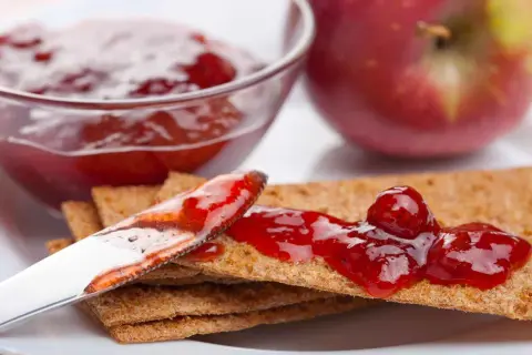 Un cucchiaio spalma la marmellata di mele su cracker croccanti, con una ciotola piena e una mela rossa sullo sfondo.