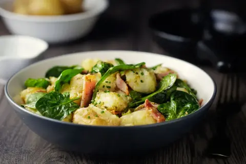 Insalata di patate con spinacini freschi e pancetta croccante, condita con erbe aromatiche. Un piatto gustoso e colorato.