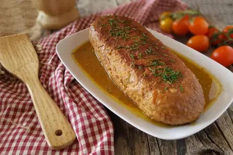 Polpettone in padella dorato e succulento, guarnito con erba cipollina, su un piatto bianco con salsa. Accanto, una spatola di legno e pomodorini.