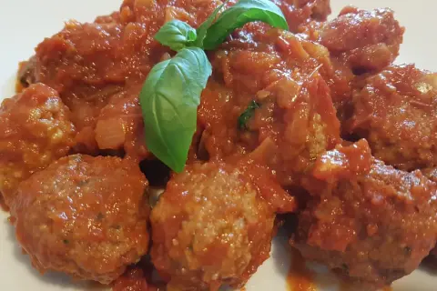 Un piatto invitante di polpette di carne morbide immerse in un sugo rosso ricco, guarnite con una foglia di basilico fresco.