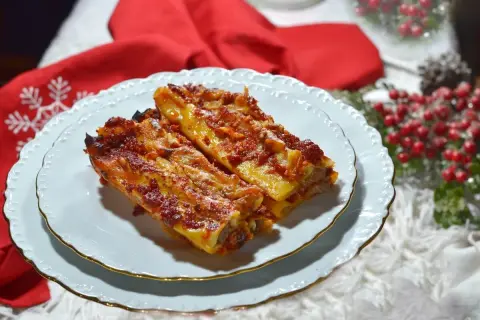 Due cannelloni di carne fumanti, ricoperti di sugo rosso e formaggio fuso, serviti su un piatto bianco con decorazioni natalizie sullo sfondo.