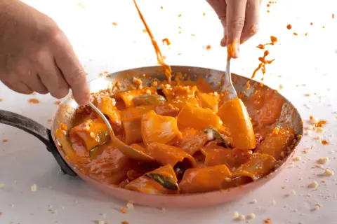 Mani che mescolano paccheri alla Vittorio in padella, con salsa arancione che schizza.