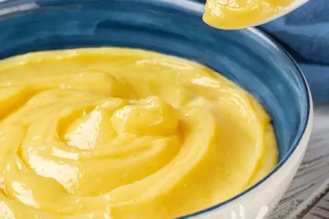 Una ciotola blu piena di vellutata crema pasticcera senza uova, con un cucchiaio che ne solleva una porzione.