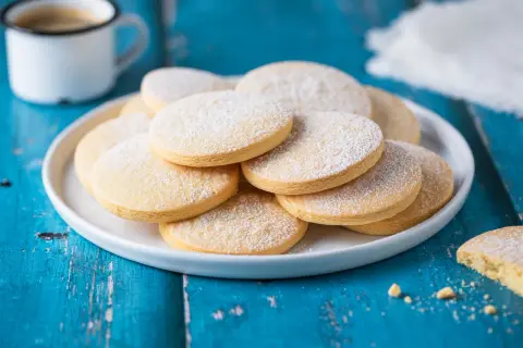 Biscotti senza lattosio spolverati di zucchero a velo su un piatto bianco, accanto a una tazza di caffè.