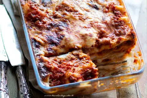 Lasagne alla bolognese appena sfornate, dorate e filanti, in una teglia di vetro. Accanto, due coltelli d'argento.