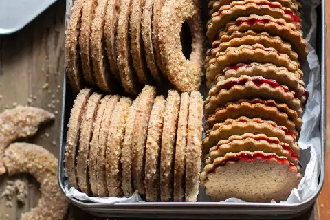 Biscotti grano saraceno ripieni di marmellata, impilati in una scatola di latta. Alcuni biscotti a forma di ciambella e a mezzaluna sono sparsi sul tavolo.