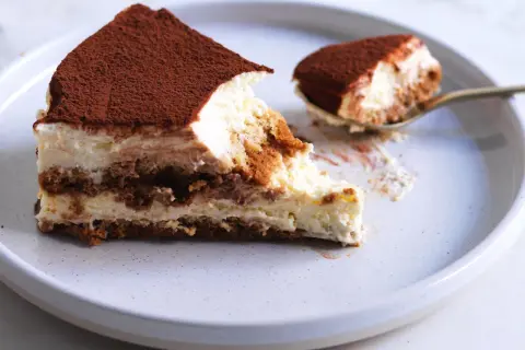 Una fetta di tiramisù ricetta originale, con strati di savoiardi, crema al mascarpone e cacao in polvere. Un cucchiaino ne ha già assaggiato un pezzetto.