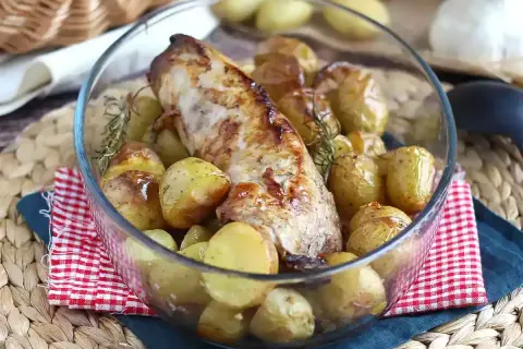 Un succulento filetto di maiale al forno, dorato e profumato, circondato da patate novelle arrosto e rametti di rosmarino in una ciotola di vetro.