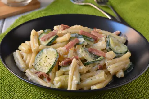 Piatto di pasta cremosa con zucchine a rondelle e cubetti di speck croccante, guarnito con pepe nero.