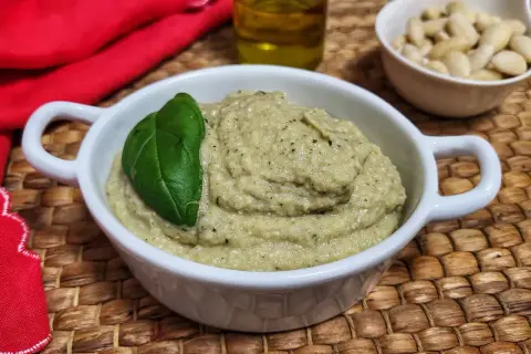 Pesto di melanzane cremoso in una ciotola bianca, guarnito con una foglia di basilico. Sullo sfondo, mandorle e olio d'oliva.