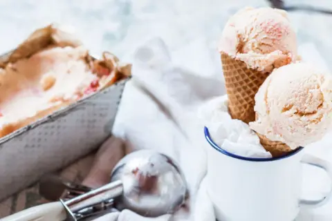 Due palline di gelato fatto in casa, con pezzetti di frutta, in un cono, accanto a una vaschetta e un porzionatore.