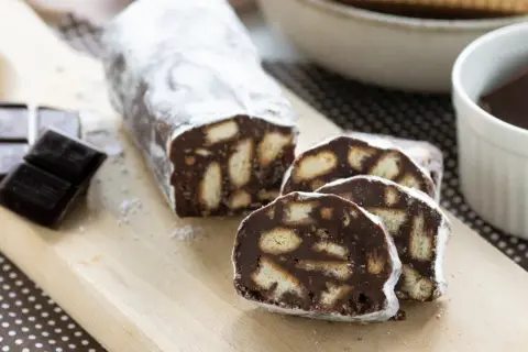 Un delizioso salame al cioccolato, cosparso di zucchero a velo, con biscotti e cioccolato fondente a lato.