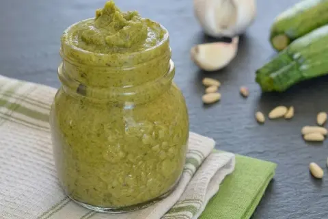 Un vasetto pieno di cremoso pesto di zucchine, con ingredienti freschi come zucchine, aglio e pinoli sullo sfondo.