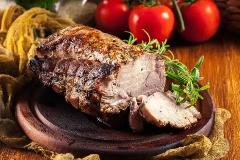 Arista di maiale succulenta e dorata, affettata su un tagliere di legno, con rosmarino e pomodori sullo sfondo.