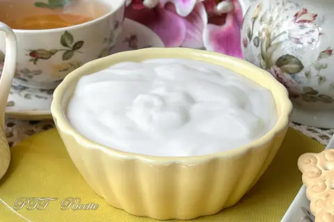 Una ciotola gialla piena di soffice crema al latte, accanto a una tazza di tè e fiori.