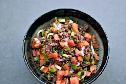 Una fresca insalata di lenticchie estiva, con pomodori, cipolla rossa e erbe aromatiche, servita in una ciotola scura.