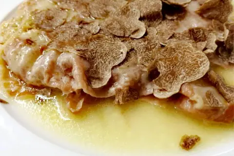 Cotoletta alla bolognese ricoperta di scaglie di tartufo, servita in un piatto bianco con una salsa dorata.