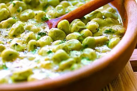 Un primo piatto saporito: piselli in umido, cremosi e profumati, serviti in una ciotola di legno con un cucchiaio.