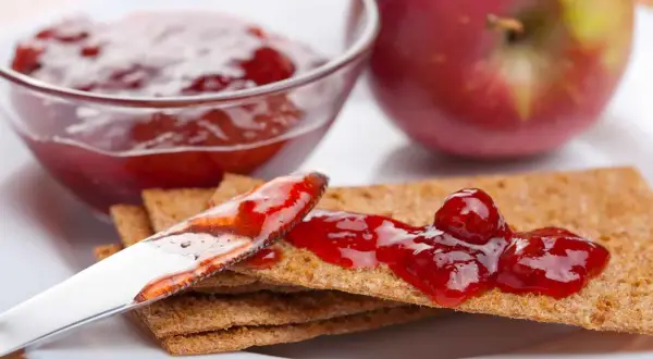 Un cucchiaio spalma la marmellata di mele su cracker croccanti, con una ciotola piena e una mela rossa sullo sfondo.
