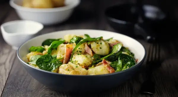 Insalata di patate con spinacini freschi e pancetta croccante, condita con erbe aromatiche. Un piatto gustoso e colorato.