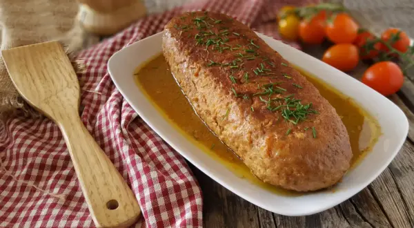 Polpettone in padella dorato e succulento, guarnito con erba cipollina, su un piatto bianco con salsa. Accanto, una spatola di legno e pomodorini.