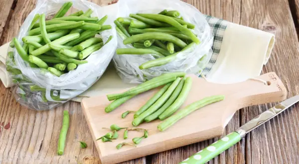 Fagiolini freschi in sacchetti trasparenti, pronti per essere preparati. Un tagliere di legno e un coltello suggeriscono come congelare i fagiolini per conservarli.