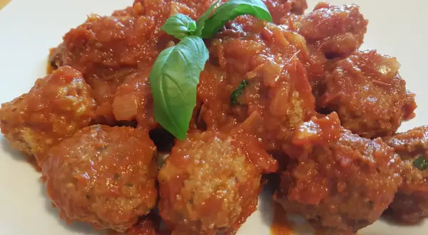 Un piatto invitante di polpette di carne morbide immerse in un sugo rosso ricco, guarnite con una foglia di basilico fresco.