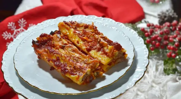 Due cannelloni di carne fumanti, ricoperti di sugo rosso e formaggio fuso, serviti su un piatto bianco con decorazioni natalizie sullo sfondo.