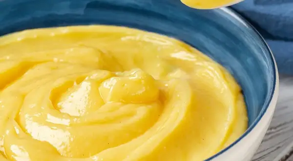 Una ciotola blu piena di vellutata crema pasticcera senza uova, con un cucchiaio che ne solleva una porzione.