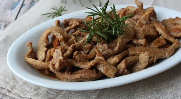 Deliziosi straccetti di vitello saltati, guarniti con rosmarino fresco, serviti in un piatto bianco su un tovagliolo di lino.