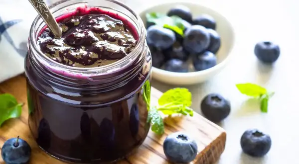 Vasetto di marmellata di mirtilli fatto in casa, con mirtilli freschi e foglie di menta su un tagliere di legno.