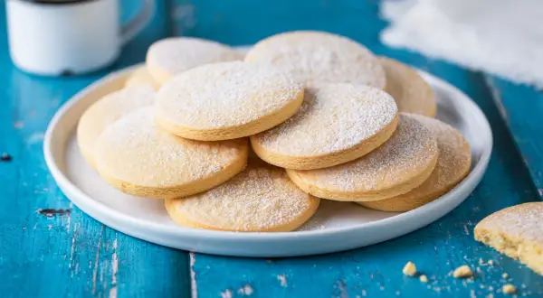 Biscotti senza lattosio spolverati di zucchero a velo su un piatto bianco, accanto a una tazza di caffè.