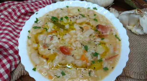 Un piatto fumante di riso e fagioli, condito con olio d'oliva e prezzemolo, con aglio e una tovaglia a scacchi sullo sfondo.